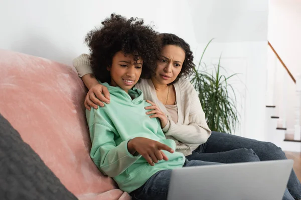 confused african american preteen girl pointing at laptop near curly mother