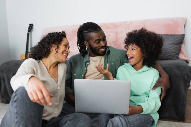 Neşeli Afro-Amerikan ailesi evde dizüstü bilgisayarın yanında gülümseyip birbirlerine bakıyorlar.