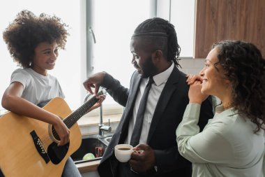 Mutlu Afro-Amerikalı bir adam elinde bir fincan kahveyle genç kızına akustik gitar çalmayı öğretiyor.