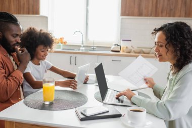 Mutfakta dijital tablet kullanan mutlu bir Afro-Amerikan kadını dizüstü bilgisayar ve ailesinin yanında kağıt tutuyor. 
