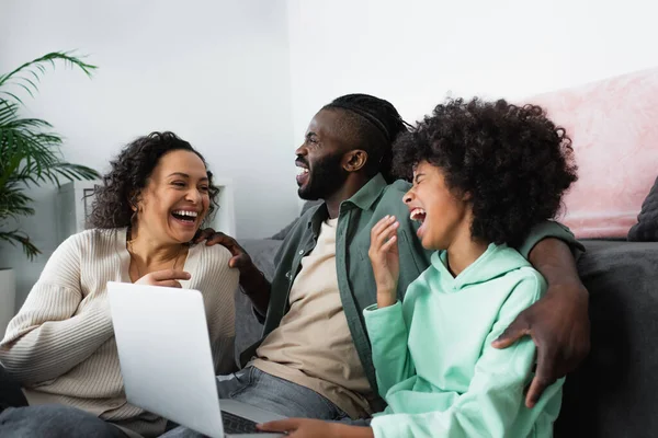 cheerful african american family laughing and watching movie on laptop at home