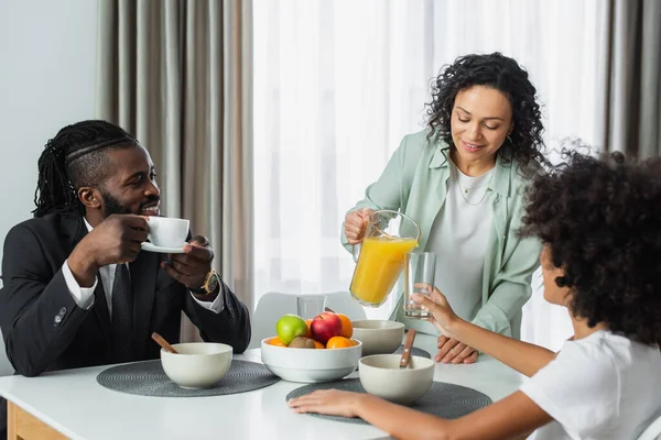 幸せなアフリカ系アメリカ人女性を注ぐオレンジジュースでガラス近く夫でスーツと十代の娘で朝食中 — ストック写真
