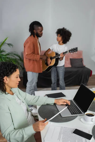 Kıvırcık Afrikalı Amerikalı kadın dijital tablet tutuyor ve kocasının yanında akustik gitar çalan kızının yanında dizüstü bilgisayar kullanıyor.