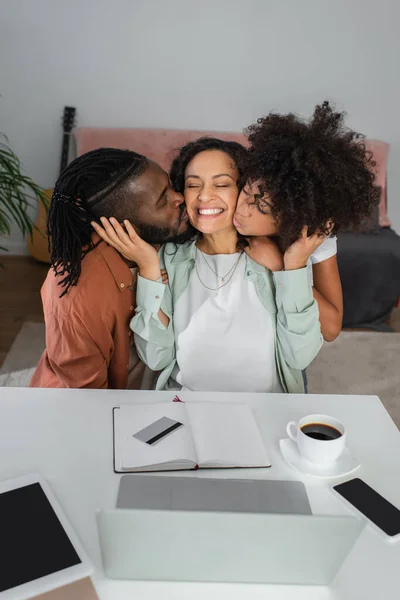 african american man and preteen girl kissing cheeks of happy woman near devices on desk