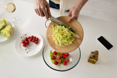 Kesilmiş adamın kaseye kıvırcık salata ve akıllı telefonun yanında boş ekran domatesli domates ekleyişinin üst görüntüsü. 