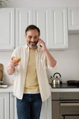 Beyaz gömlekli neşeli adam mutfakta bir bardak şarapla dururken cep telefonuyla konuşuyor.