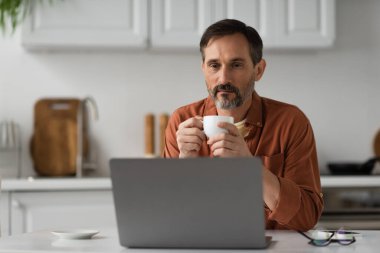 Kahve fincanı tutan ve mutfaktaki laptopa bakan sakallı adam.