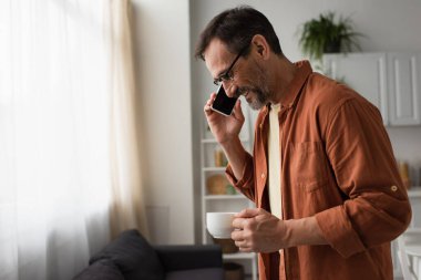 Mutfaktaki akıllı telefondan sabah kahvesi içen gözlüklü gülümseyen adamın yan görüntüsü.