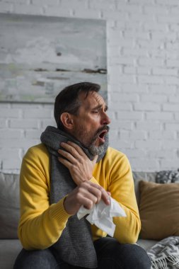 sick man in warm scarf holding paper napkin and coughing on couch clipart