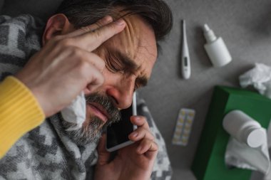 Yüksek ateş ve baş ağrısı çeken hasta bir adamın ilaç ve konteynırların yakınındaki akıllı telefondan tedavi gördüğü görülüyor.