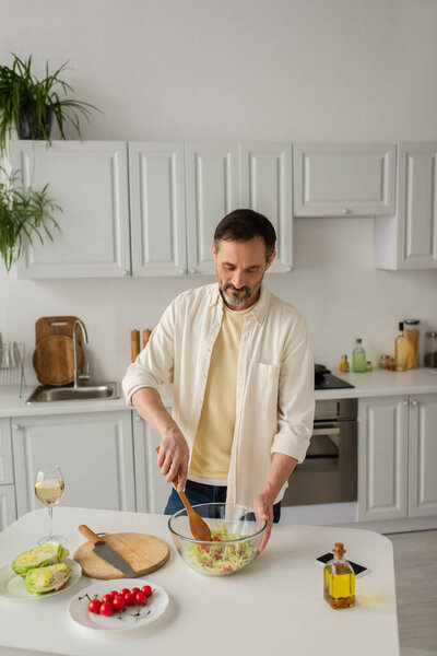 man in white shirt preparing vegetable salad with lettuce and cherry tomatoes near glass of wine