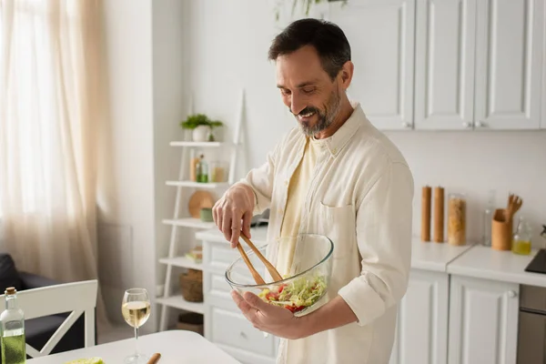 明るい男は台所で白ワインのガラスの近くの木のトングと野菜サラダを混合 — ストック写真