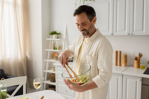 cheerful man mixing vegetable salad with wooden tongs near glass of white wine in kitchen