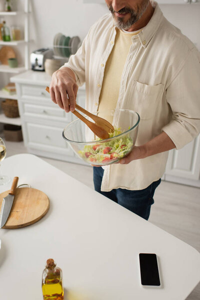 partial view of smiling man mixing fresh vegetable salad with wooden tongs while cooking in kitchen