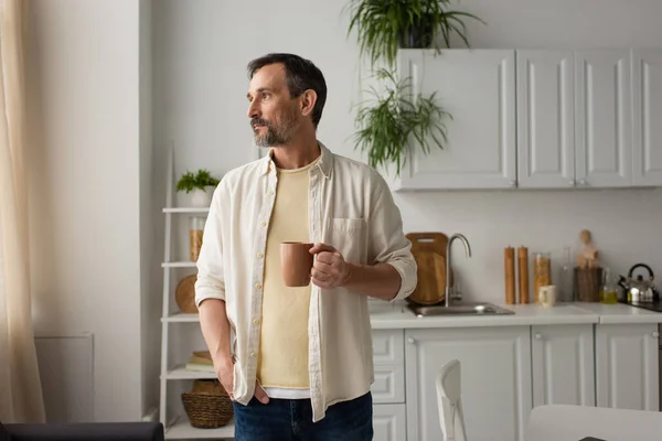 bearded man in white shirt standing with cup of tea and hand in pocket while looking away in kitchen