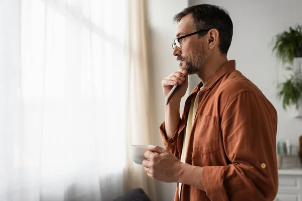 side view of thoughtful man in eyeglasses standing with smartphone and coffee cup near window at home
