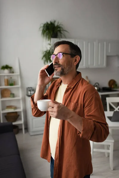 Hombre Barbudo Anteojos Hablando Teléfono Inteligente Sosteniendo Taza Café Cocina — Foto de Stock