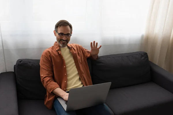 joyful bearded man waving hand during video call on laptop while sitting on sofa