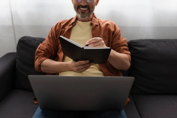 幸福的男人坐在沙发上 拿着笔记本电脑 在笔记本上写字的部分画面 — 图库照片