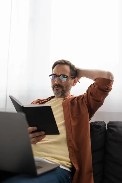 Nachdenklicher Mann Mit Brille Schaut Notizbuch Während Mit Verschwommenem Laptop — Stockfoto