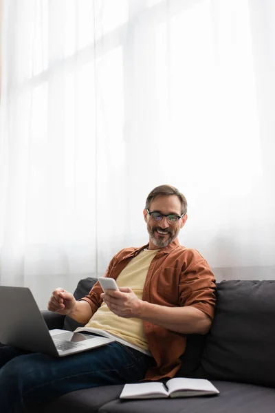 Leende Skäggig Man Håller Smartphone Och Tittar Tom Anteckningsbok Medan — Stockfoto