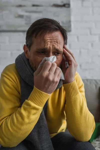 ill man in warm scarf sneezing in paper napkin and suffering from migraine at home