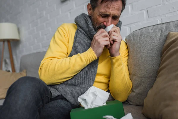 病気の男で暖かいスカーフくしゃみでペーパーナプキンとともに閉じた目でソファ — ストック写真