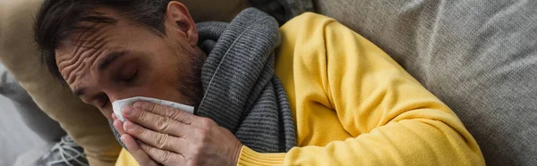 ソファに寝そべって目を閉じたままくしゃみをする紙ナプキンを持つ病気の男のトップビュー バナー — ストック写真