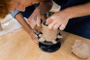 Blurred man forming clay cup near african american teacher in pottery workshop  clipart