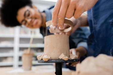 Man holding clay near sculpture and blurred african american teacher in pottery workshop  clipart