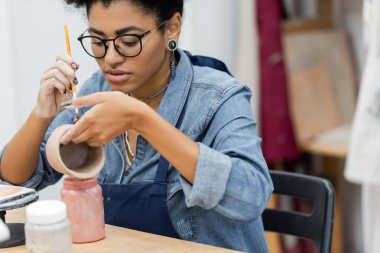 African american craftswoman in eyeglasses painting on ceramic product near jars with paint in pottery studio  clipart