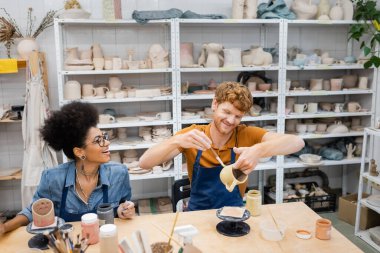 Smiling redhead man painting on clay cup near african american girlfriend in apron in pottery workshop  clipart