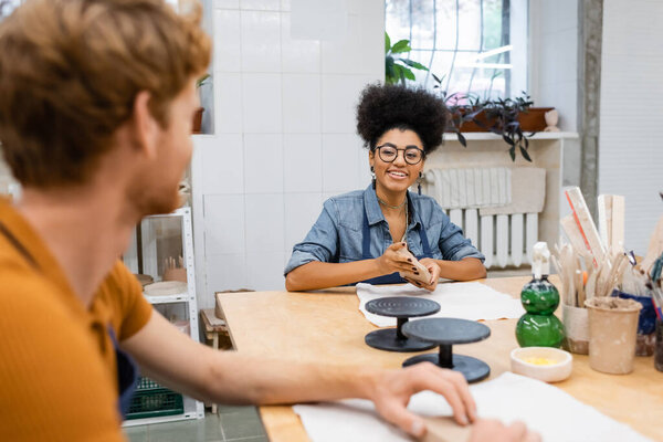 happy african american woman in eyeglasses molding and looking at redhead man on blurred foreground