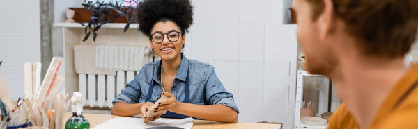 happy african american woman in eyeglasses molding and looking at redhead man on blurred foreground, banner