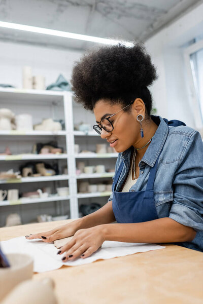 curly african american woman in apron and eyeglasses shaping clay piece with hands in pottery workshop