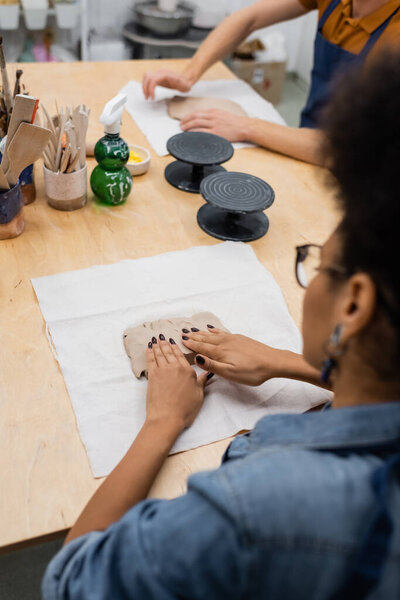 high angle view of african american woman in eyeglasses shaping clay piece with hands during pottery class