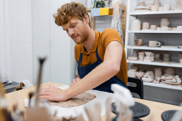 bearded redhead man in apron shaping clay piece with hand in pottery workshop