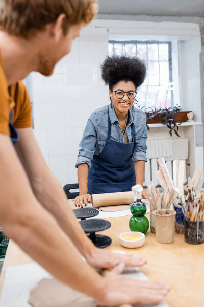 happy african american woman in eyeglasses shaping clay piece with rolling pin near redhead man on blurred foreground