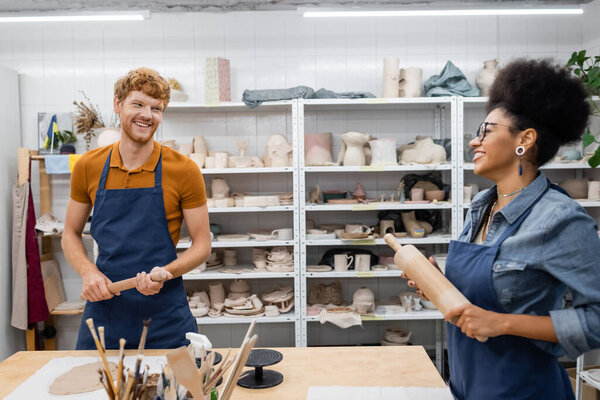 happy interracial couple in aprons holding rolling pins while looking at each other during pottery class