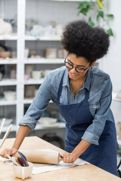 happy african american woman in eyeglasses shaping clay piece with rolling pin