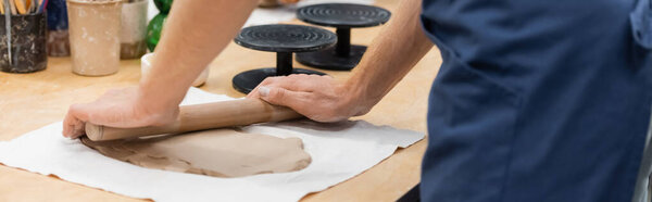 cropped view of man in apron modeling clay piece with rolling pin, banner