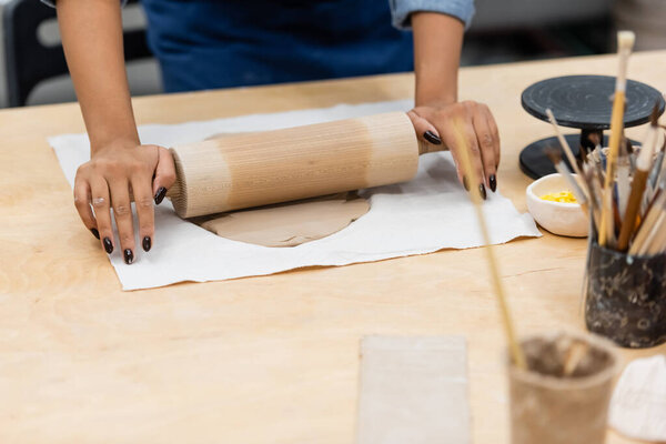 partial view of young african american woman in apron modeling clay with rolling pin near paintbrushes 