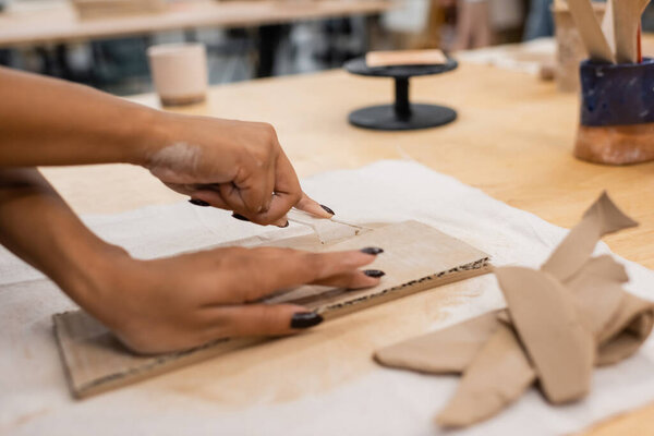 partial view of african american woman cutting clay with knife while handcrafting in pottery workshop
