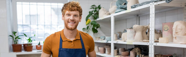 positive redhead man in apron smiling near sculptures on shelves of rack in pottery shop, banner