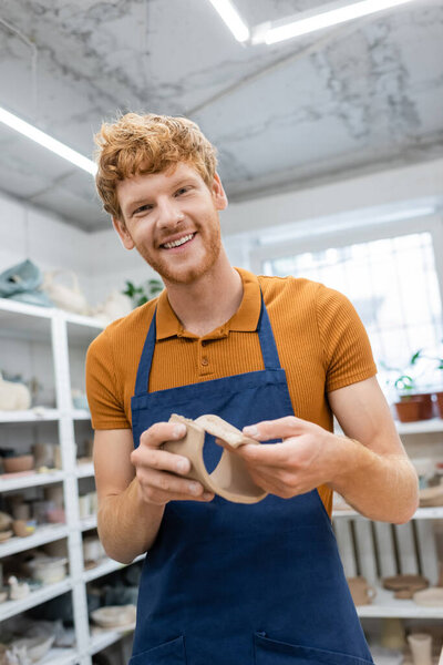cheerful man with red hair standing in apron and modeling clay piece during pottery class