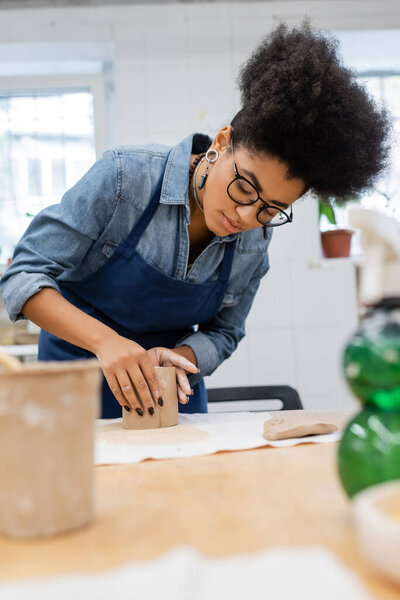 young african american woman in apron modeling clay piece during pottery class 