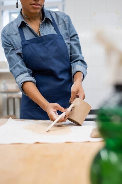 partial view african american woman in apron holding shaper while modeling clay in pottery workshop