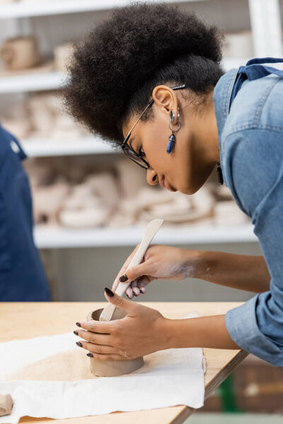 curly african american woman in eyeglasses holding shaper while modeling clay cup during pottery class 