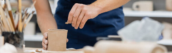 cropped view of young man in apron shaping clay piece into cup, banner