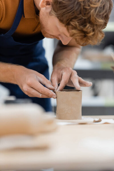 curly and bearded man with red hair shaping clay piece into cup 